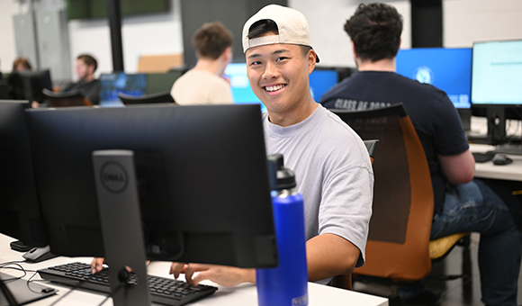 A student with a backwards white hat smiles from behind a computer.