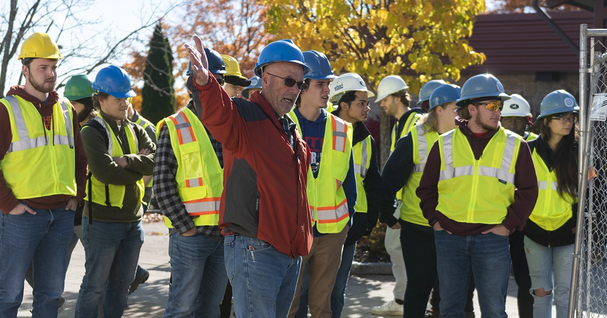 A site manager leads a group of students dressed in reflective yellow vests and hard hats at a construction site.