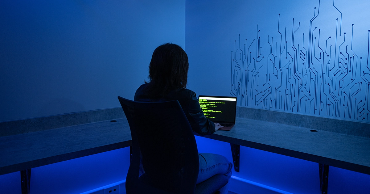 A hacker works from a small room lit in blue light.