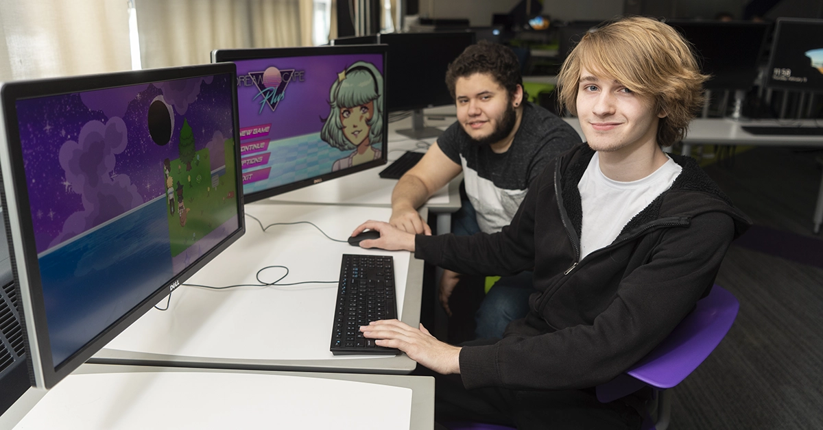 Two Game Design students sit next to computers displaying the landing screen of their game, Dream Cafe Plus.