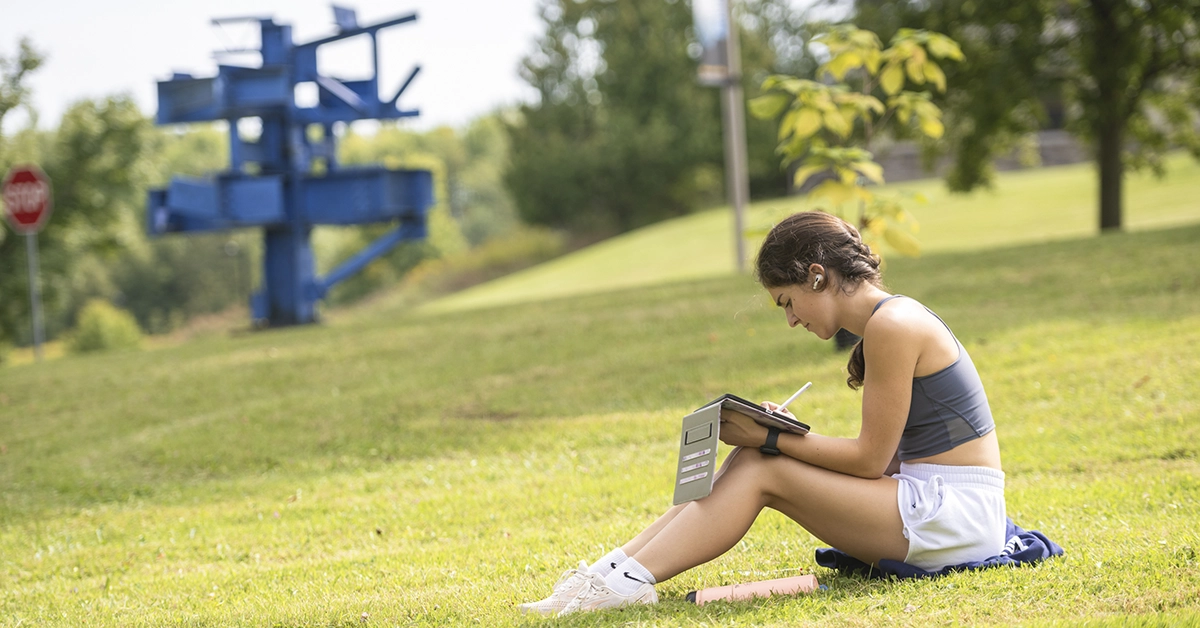 A student sits on the grass and sketches on an iPad outside Nevaldine Hall. A large blue sculpture sits in the background.