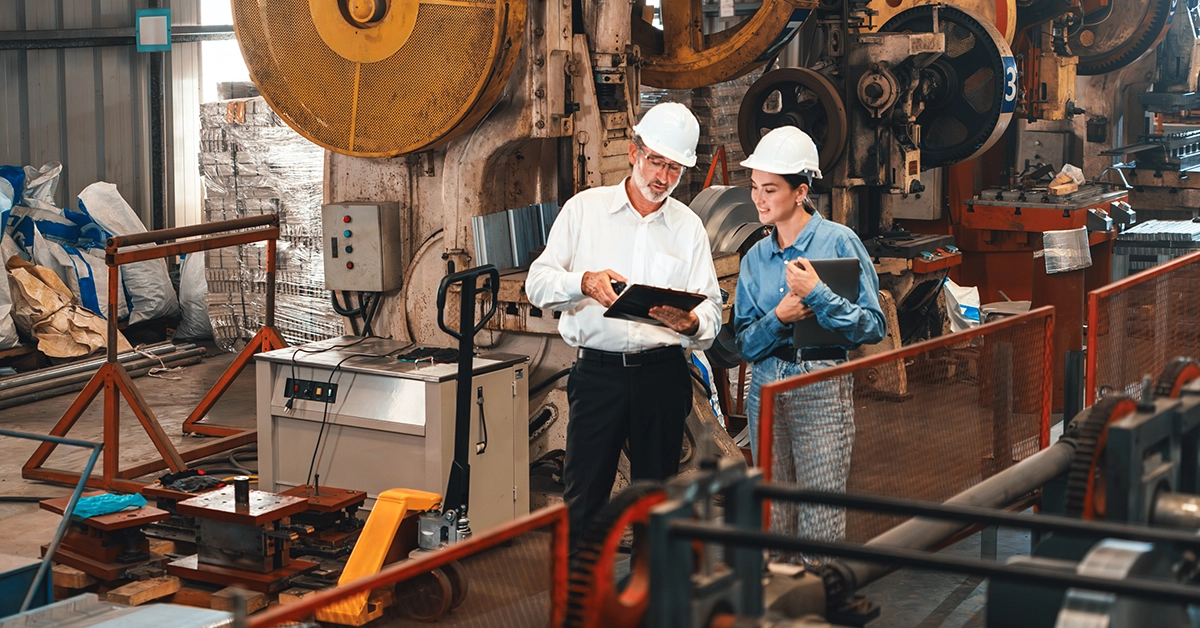 Two manufacturing employees wearing white hard hats review notes from a clipboard.