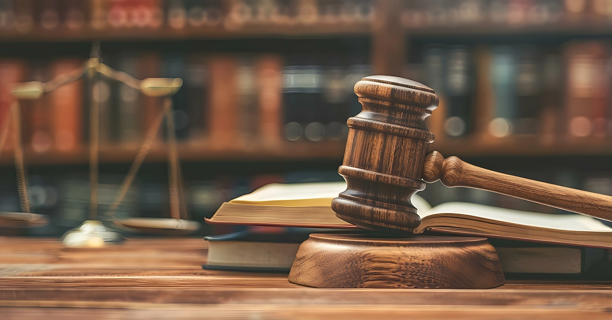 A gavel sits on a table in a room with legal books and the scales of justice.